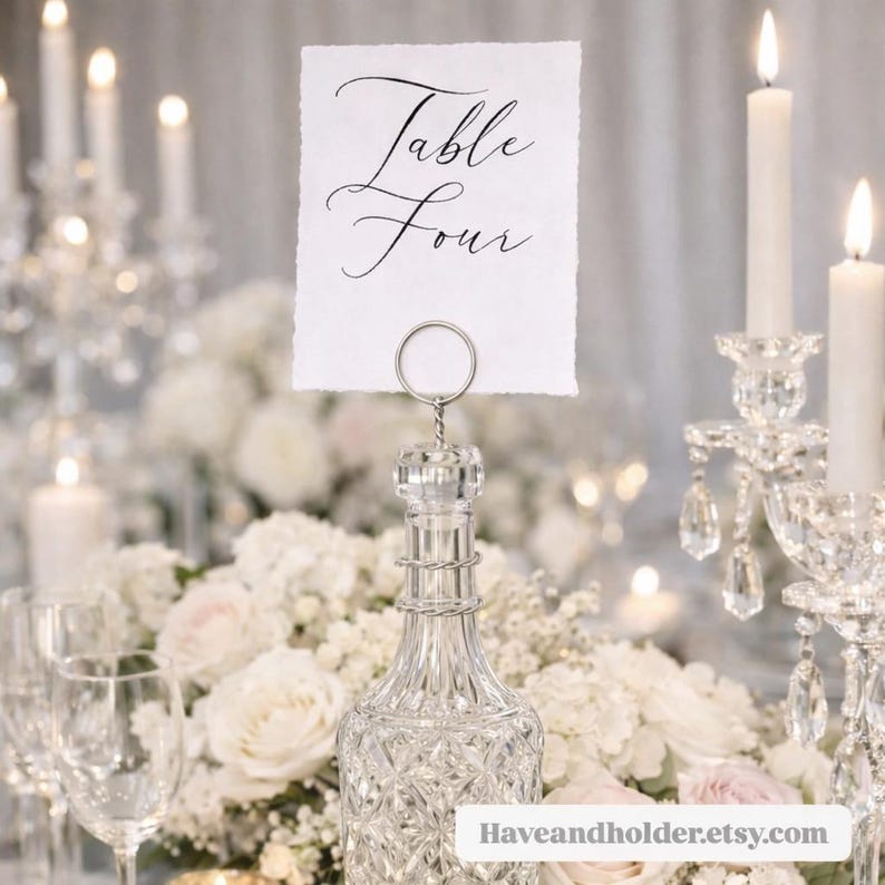 May include: A clear glass bottle with a decorative stopper holds a white card that reads "Table Four" in elegant script. The bottle is surrounded by white flowers, candles, and crystal accents, creating a sophisticated table setting.
