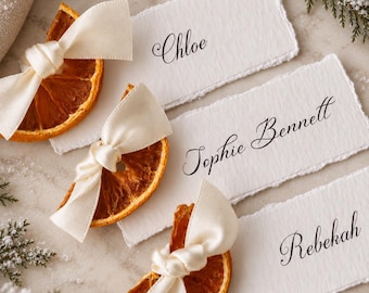 May include: Three rectangular white place cards with deckled edges, each featuring a black printed name: Chloe, Sophie Bennett, and Rebekah. Each card is paired with a dried orange slice adorned with a tied cream-colored satin ribbon.