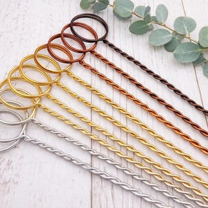 May include: A selection of metal stirrers in silver, gold, copper, and brown. Each stirrer has a twisted stem and a circular handle. The stirrers are displayed on a light wood surface.