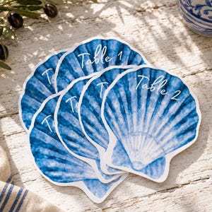 Op de afbeelding: Een stapel uitgestanste papieren tafelkaartjes in de vorm van schelpen. Elke kaart heeft een blauw-witte aquarelprint van een sint-jakobsschelp. De tekst "Table 1" en "Table 2" is in witte schrijfletters op de kaarten gedrukt.