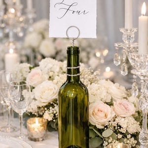Può includere: Una bottiglia di vino verde usata come portanumero da tavolo, con un dettaglio in filo d'argento attorno al collo. Un biglietto bianco con le parole "Table Four" è tenuto in posizione. L'allestimento del tavolo comprende candele, fiori e vetreria.