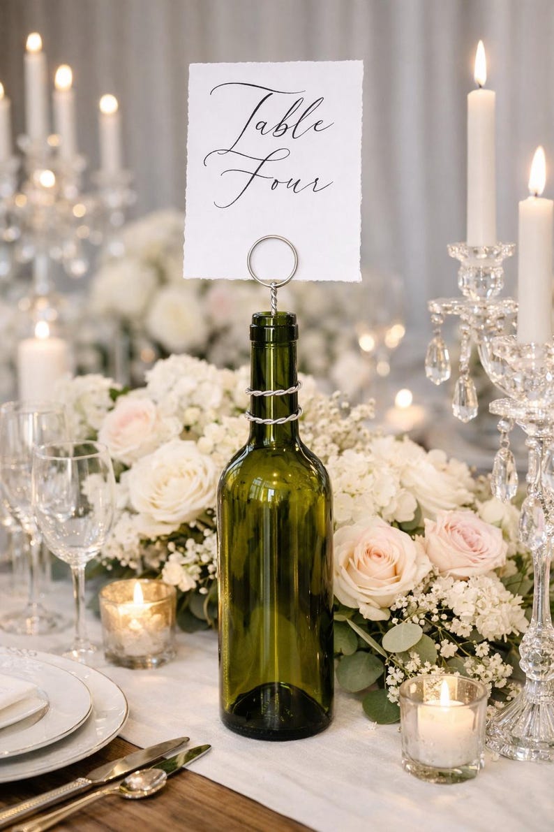 May include: A green wine bottle used as a table number holder, with a card that reads "Table Four" in elegant script. The bottle is decorated with a silver wire. The table setting includes candles, flowers, and glassware.