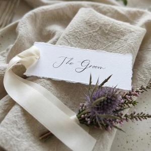 May include: A rectangular white paper place card with deckled edges featuring the black handwritten text The Groom. The card is tied with a cream-coloured silk ribbon and decorated with a small sprig of purple thistle and pink heather flowers.