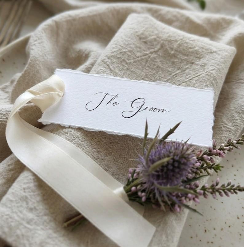 May include: A rectangular white paper place card with deckled edges featuring the black handwritten text The Groom. The card is tied with a cream-coloured silk ribbon and decorated with a small sprig of purple thistle and pink heather flowers.