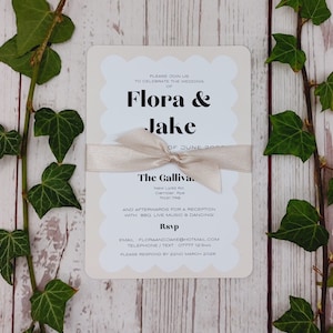 Może przedstawiać: Kremowe zaproszenie ślubne z falistą krawędzią, z imionami "Flora & Jake" pogrubioną czcionką, związane beżową wstążką. Zaproszenie zawiera szczegóły ślubu, z zielonym bluszczem na drewnianym tle.