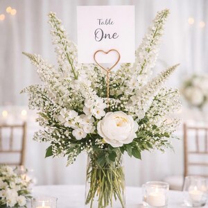 May include: A clear glass vase filled with white flowers including tall delphiniums and a large central peony. A rose gold heart-shaped wire holder displays a white card with the black text Table One centered above the floral arrangement.