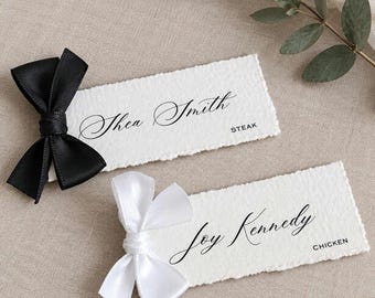 May include: Two rectangular place cards with deckled edges, one featuring a black satin bow and the name Shea Smith with the text STEAK, and the other with a white satin bow and the name Joy Kennedy with the text CHICKEN.