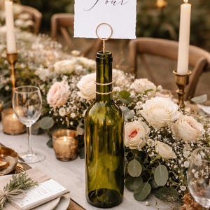 May include: A table setting with a green wine bottle used as a table number holder. The card reads "Table Four" in elegant script. The table is decorated with flowers, candles, and gold accents, creating a romantic ambiance.