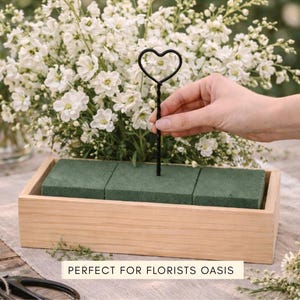 May include: A rectangular light-colored wooden box containing three green floral foam blocks. A black metal wire stake with a heart-shaped top is inserted into the center foam block. The text "PERFECT FOR FLORISTS OASIS" is printed on a label.