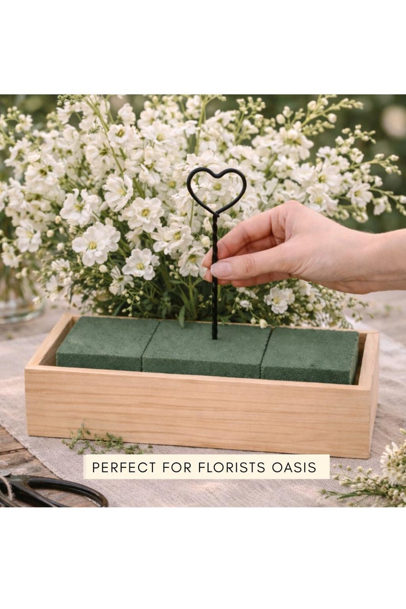 May include: A rectangular light-colored wooden box containing three green floral foam blocks. A black metal wire stake with a heart-shaped top is inserted into the center foam block. The text "PERFECT FOR FLORISTS OASIS" is printed on a label.