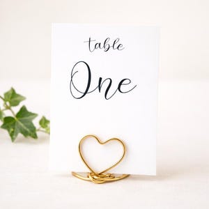 Puede incluir: Una tarjeta de mesa blanca con la palabra "table" y "One" en elegante escritura negra. La tarjeta está sujeta por un soporte de alambre dorado en forma de corazón, ideal para bodas o eventos.