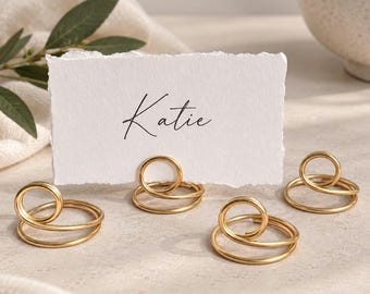 Pode incluir: Três suportes para cartões de lugar em fio de metal dourado com um design de espiral circular, cada um com uma base plana e um laço vertical para prender um cartão de papel branco retangular com bordas irregulares e o nome Katie impresso em preto.