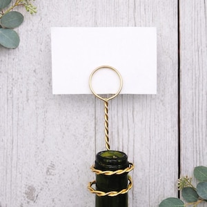May include: A gold-colored place card holder with a twisted stem, holding a white card. The holder is attached to a dark green bottle, wrapped with gold wire. The background is a light wood surface.