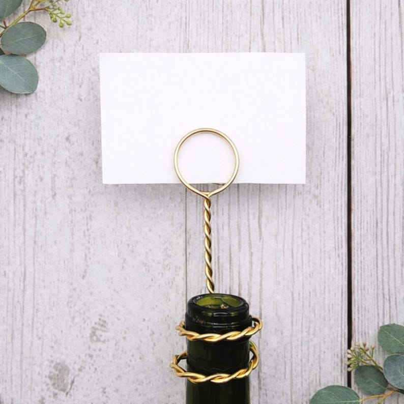 May include: A gold-colored place card holder with a twisted stem, holding a white card. The holder is attached to a dark green bottle, wrapped with gold wire. The background is a light wood surface.