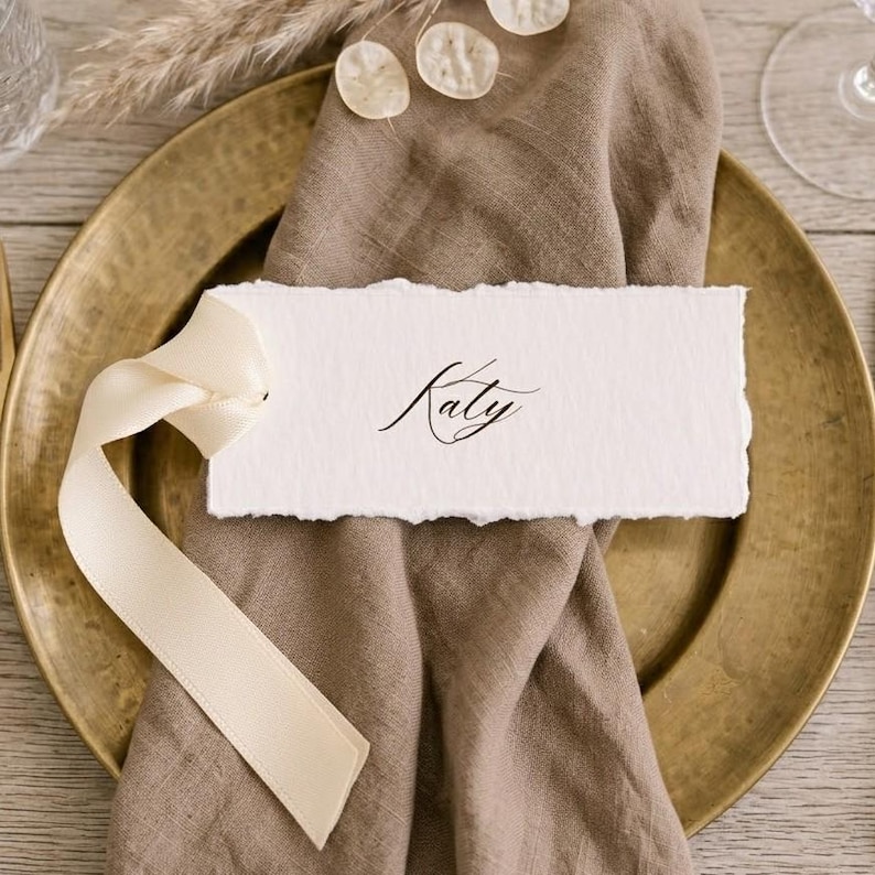 May include: A rectangular white place card with deckled edges featuring the name Katy in black cursive script. The card is tied with a cream-coloured satin ribbon and rests on a folded brown linen napkin placed atop a round brass-toned plate.