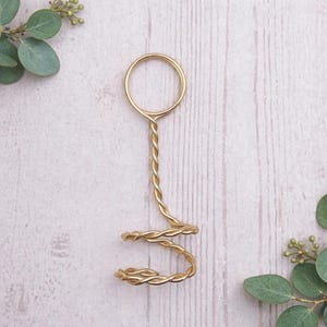 May include: A gold-colored, twisted wire ring holder with a circular top and a spiral base. The holder is designed to hold a ring. The background is a light wood grain surface with green foliage.