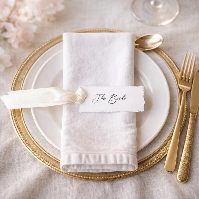 May include: A white cloth napkin folded on a white plate, featuring a cream-coloured ribbon tied around it. A rectangular white paper place card with torn edges is attached to the ribbon, displaying the black handwritten text The Bride.