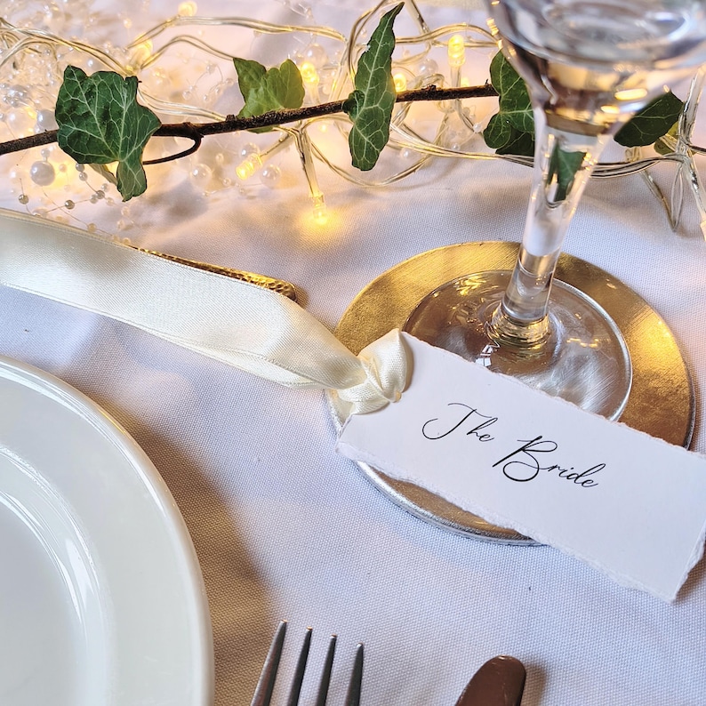 May include: A white paper place card with the text The Bride written in black script, attached to a cream-coloured satin ribbon. The card rests on a gold-coloured circular coaster beneath the base of a clear glass stemware piece.