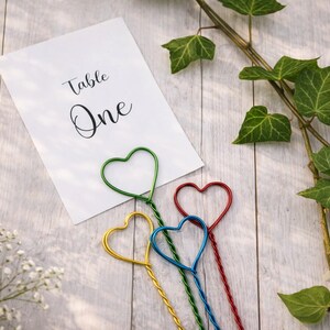 May include: A white rectangular card with the black printed text Table One, accompanied by four metal wire holders shaped like hearts in green, yellow, blue, and red. The wire stems are twisted and extend downward from the heart-shaped tops.