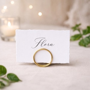 Könnte beinhalten: Eine weiße Tischkarte mit dem Namen "Flora" in eleganter Schrift, gehalten von einem goldenen Ringhalter. Die Karte hat einen Büttenrand. Der Hintergrund umfasst eine brennende Kerze und weiche, neutrale Töne.