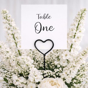 Op de afbeelding: Een witte kaart met de woorden "Table One" in zwart schrift wordt vastgehouden door een zwarte hartvormige draadkaarthouder. De kaart en de houder zijn omgeven door een weelderig arrangement van witte bloemen, wat een elegant middelpunt creëert.