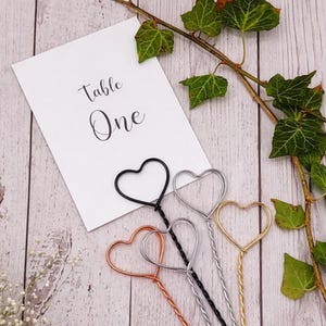 May include: Four metal wire place card holders with heart-shaped tops in black, silver, copper, and gold finishes. A white rectangular card with the black printed text "Table One" is positioned behind the wire holders.