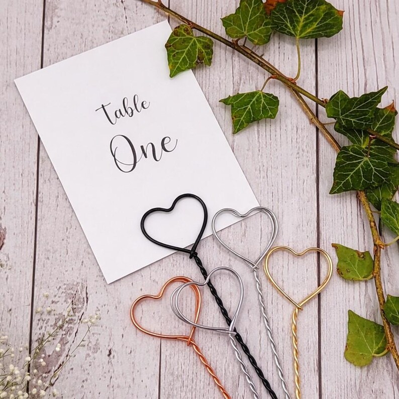 May include: Four metal wire place card holders with heart-shaped tops in black, silver, copper, and gold finishes. A white rectangular card with the black printed text "Table One" is positioned behind the wire holders.