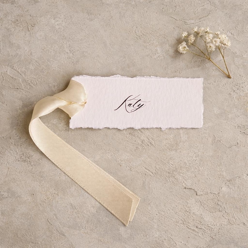 May include: A rectangular white paper tag with deckled edges featuring the name Katy written in black cursive script. A long, smooth, cream-coloured satin ribbon is tied through a hole on the left side, and a small sprig of white baby's breath flowers.