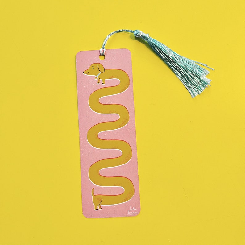 May include: A pink bookmark with a yellow dachshund illustration. The dachshund is curled into a long, winding shape. The bookmark has a light blue tassel.