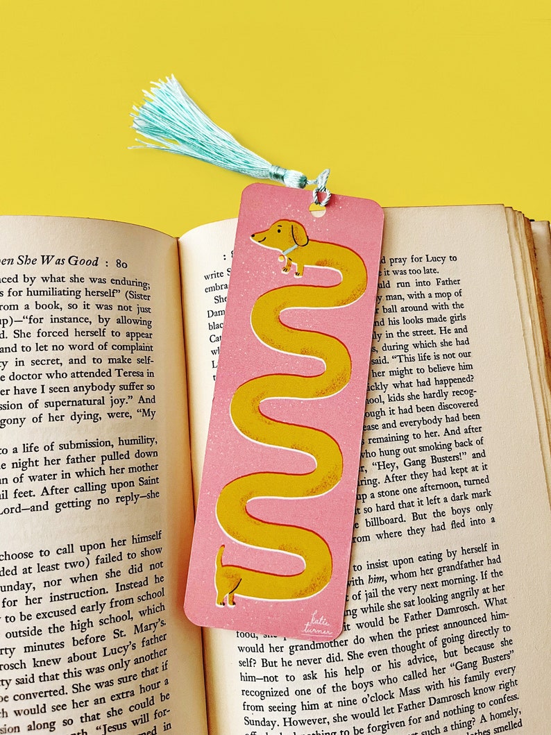 May include: A pink bookmark with a yellow dachshund illustration. The bookmark has a light blue tassel.