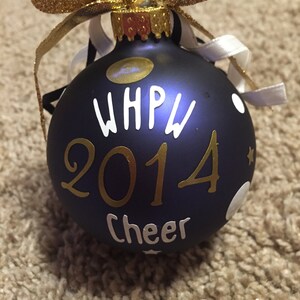 May include: A navy blue Christmas bauble with gold and white lettering and accents. The bauble has the text "WHPW 2014 Cheer" and is decorated with white polka dots and a gold glitter bow.