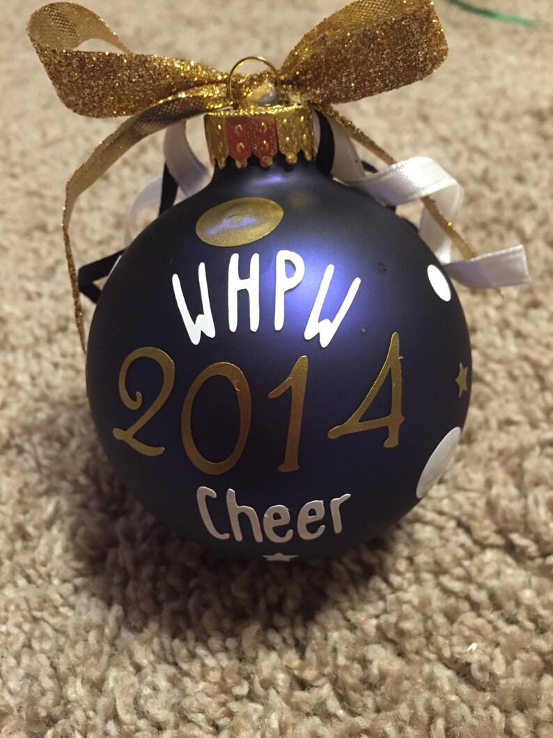 May include: A navy blue Christmas bauble with gold and white lettering and accents. The bauble has the text "WHPW 2014 Cheer" and is decorated with white polka dots and a gold glitter bow.
