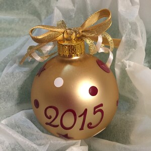 May include: A gold Christmas bauble with the year "2015" in burgundy script. The bauble has burgundy polka dots and stars, and a white dot. It is topped with a gold bow and ribbon.