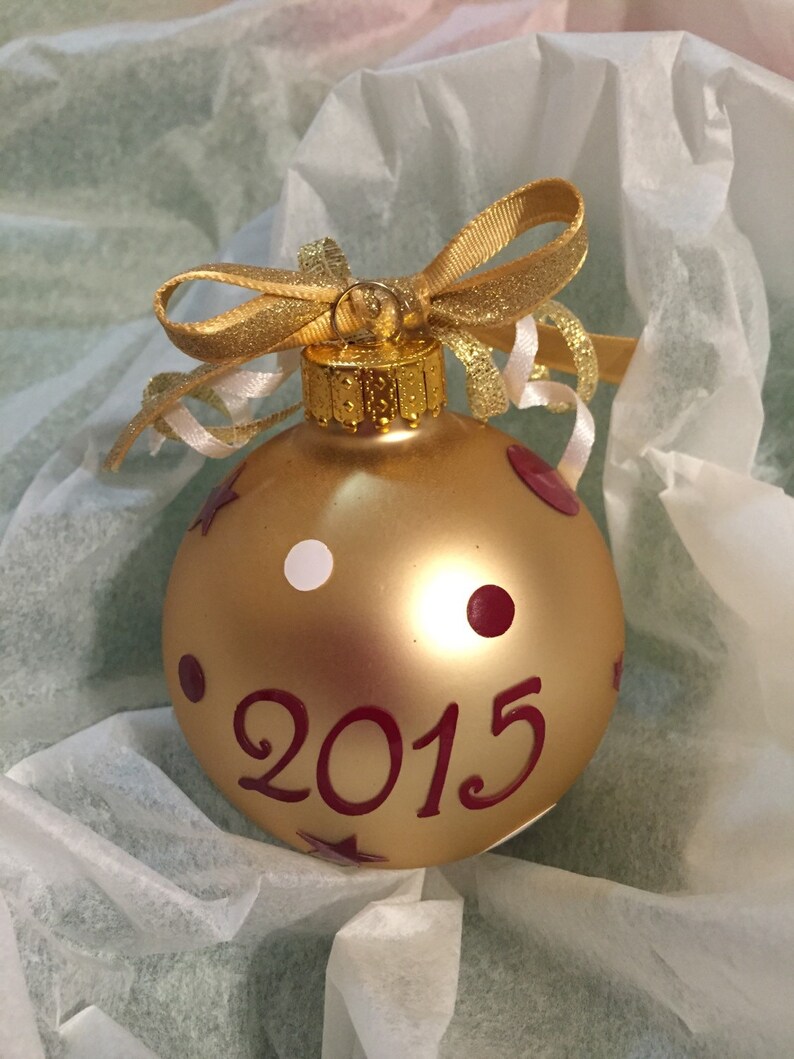 May include: A gold Christmas bauble with the year "2015" in burgundy script. The bauble has burgundy polka dots and stars, and a white dot. It is topped with a gold bow and ribbon.
