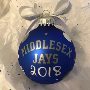 May include: A blue Christmas bauble with the text "MIDDLESEX JAYS 2018" in silver. It features white polka dots and stars, and is finished with a silver ribbon bow.