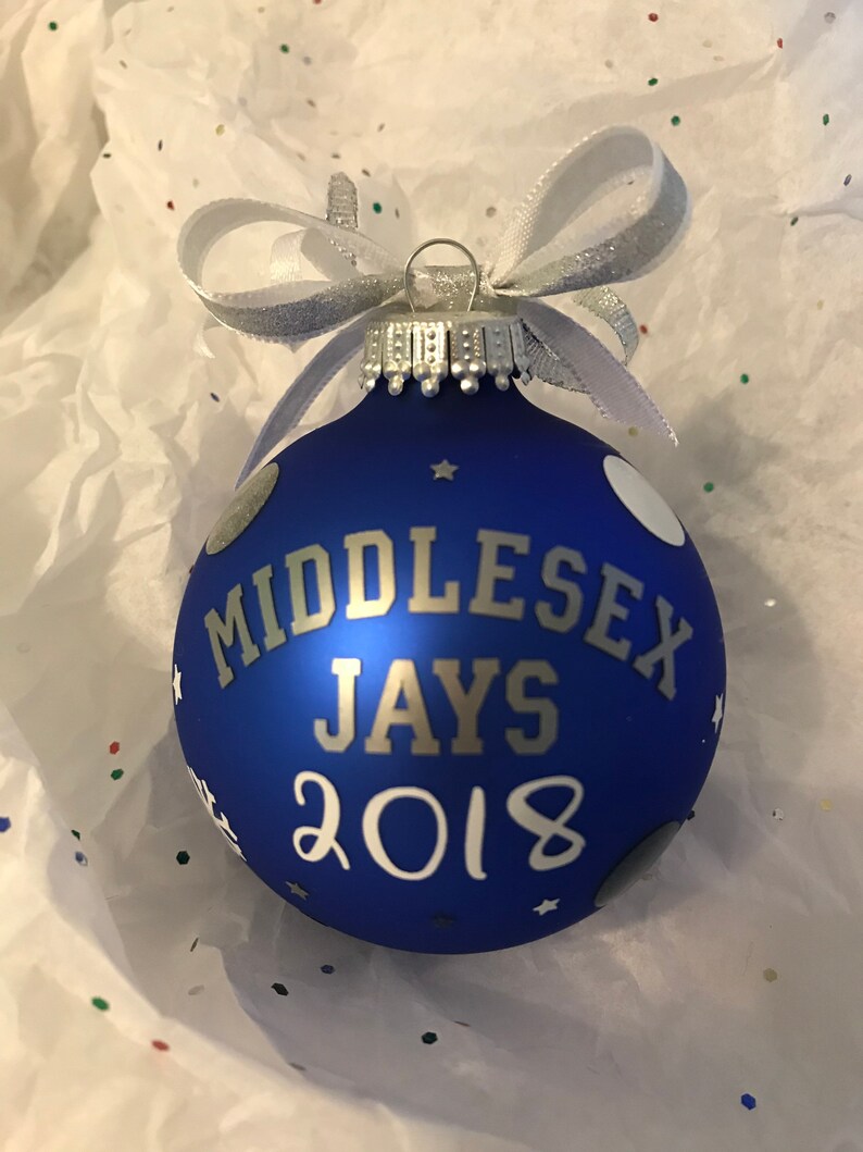 May include: A blue Christmas bauble with the text "MIDDLESEX JAYS 2018" in silver. It features white polka dots and stars, and is finished with a silver ribbon bow.