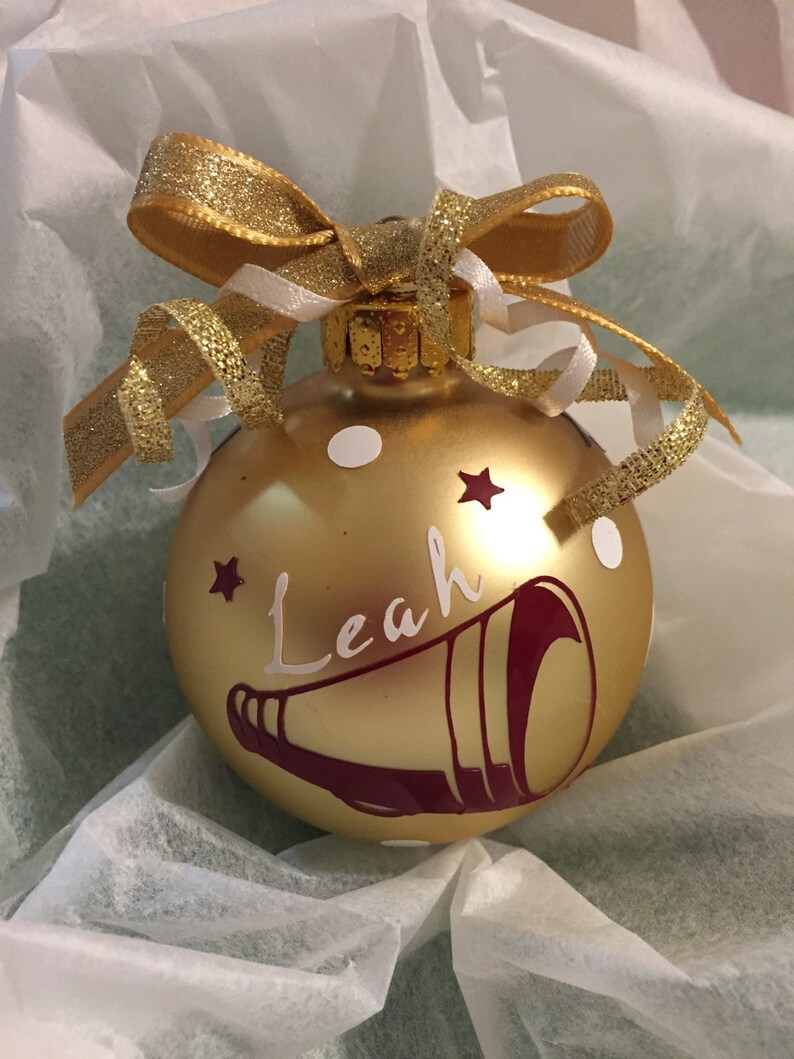 May include: A gold Christmas bauble with the name "Leah" in white script, a maroon megaphone design, and white polka dots. It is adorned with a gold glitter bow and white ribbon.