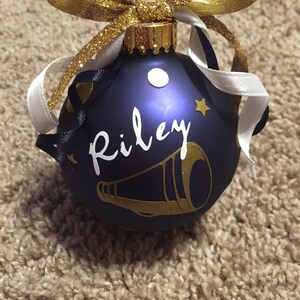 May include: A navy blue Christmas bauble with the name "Riley" in white script and a gold megaphone design. The bauble is decorated with gold and white ribbons and a gold glitter bow.