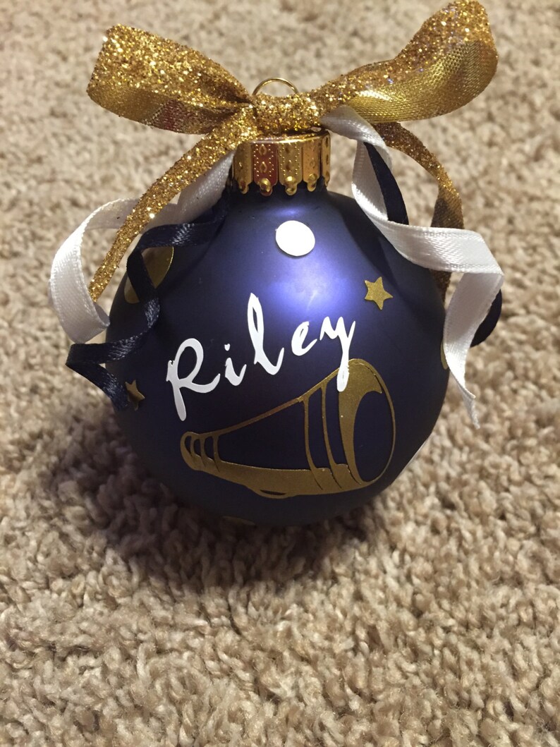 May include: A navy blue Christmas bauble with the name "Riley" in white script and a gold megaphone design. The bauble is decorated with gold and white ribbons and a gold glitter bow.
