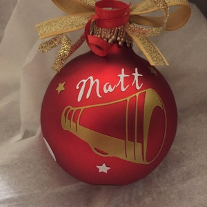 May include: Red Christmas bauble with the name "Matt" in white script and a gold megaphone design. The bauble is decorated with a gold ribbon bow and red ribbon. Small gold stars are also featured on the bauble.