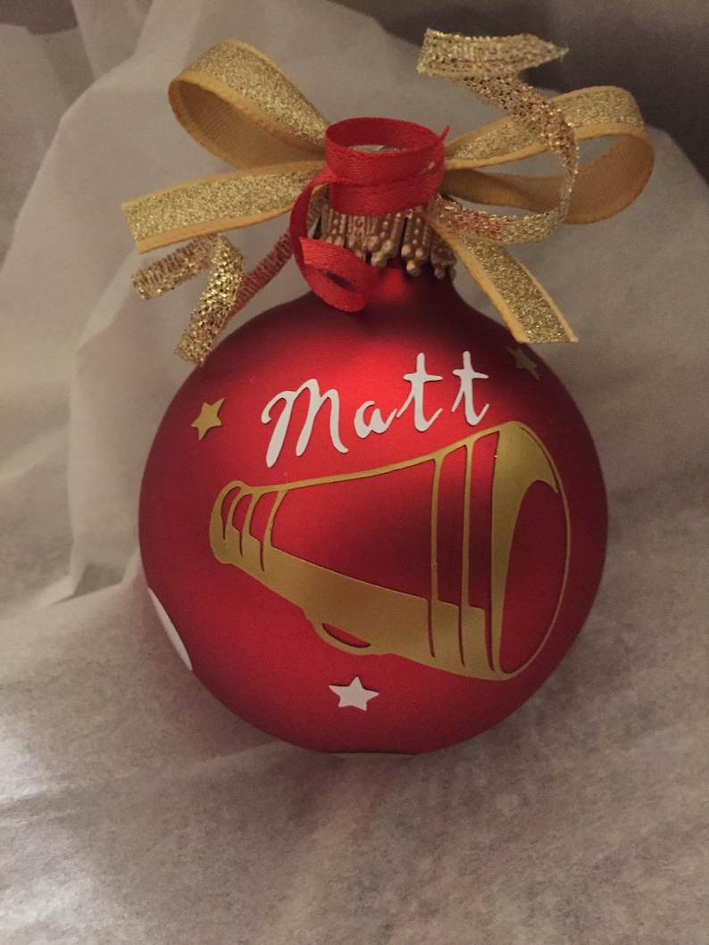 May include: Red Christmas bauble with the name "Matt" in white script and a gold megaphone design. The bauble is decorated with a gold ribbon bow and red ribbon. Small gold stars are also featured on the bauble.
