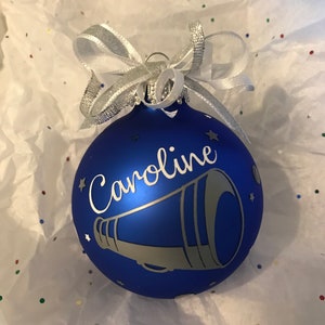 May include: A blue Christmas bauble with the name "Caroline" and a megaphone design in silver. The bauble is decorated with small star accents and a silver ribbon bow. The bauble is sitting on white tissue paper.