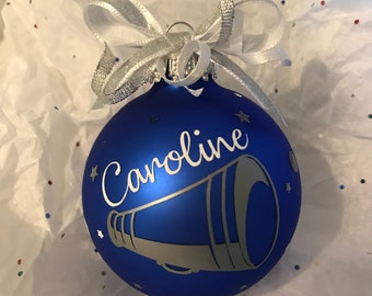 May include: A blue Christmas ornament with the name "Caroline" and a megaphone design in silver. The ornament is decorated with small star accents and a silver ribbon bow. The ornament is sitting on white tissue paper.