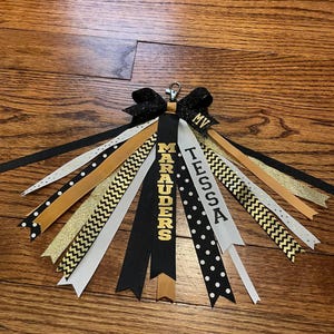 May include: A handmade black, gold, and white ribbon tassel with the word "Marauders" and the name "Tessa" printed on the ribbons. The tassel has a black bow at the top and is attached to a metal clip.