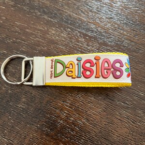 May include: Yellow keychain with a white ribbon that says "Girl Scout Daisies" in colorful letters. The ribbon has a yellow border and a small daisy flower design.