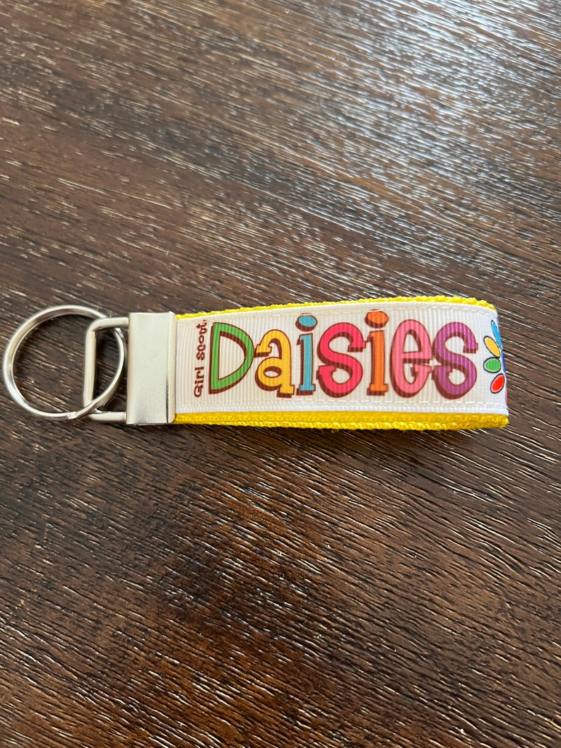 May include: Yellow keychain with a white ribbon that says "Girl Scout Daisies" in colorful letters. The ribbon has a yellow border and a small daisy flower design.