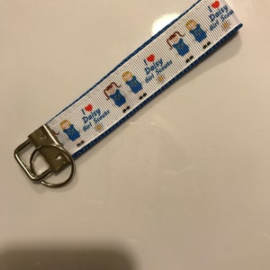 May include: A white and blue key chain with a blue ribbon and a repeating pattern of a girl scout in a blue uniform with a red heart and the text "I love Daisy Girl Scouts".