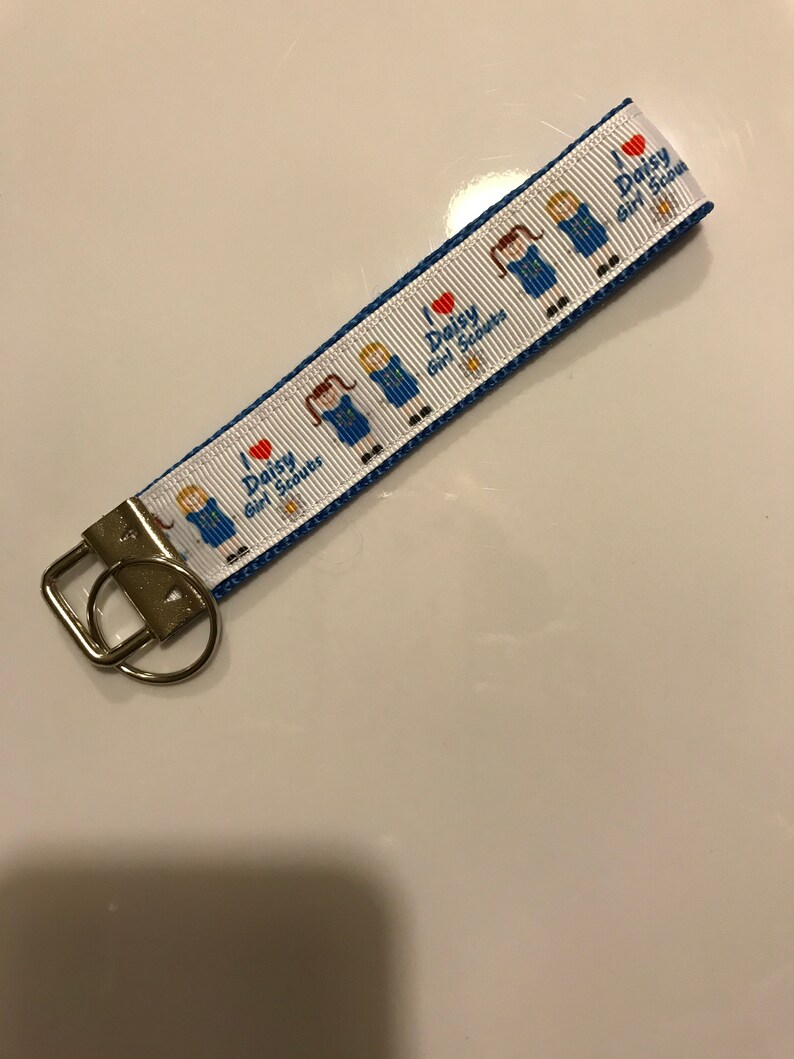 May include: A white and blue key chain with a blue ribbon and a repeating pattern of a girl scout in a blue uniform with a red heart and the text "I love Daisy Girl Scouts".