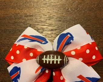 May include: A red, white, and blue bow with a football-shaped button in the center. The bow is made of ribbon with a polka dot pattern and a buffalo logo.