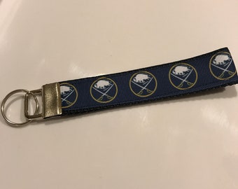 May include: A blue and gold key chain with a repeating pattern of the Buffalo Sabres logo. The key chain has a silver key ring.